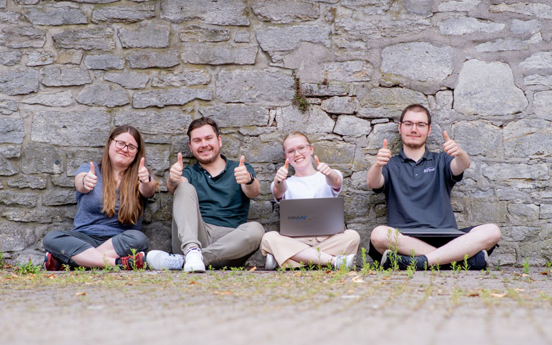 4 junge Kolleg:innen der Arineo, sitzend vor einer Mauer mit Laptop auf dem Schoß.