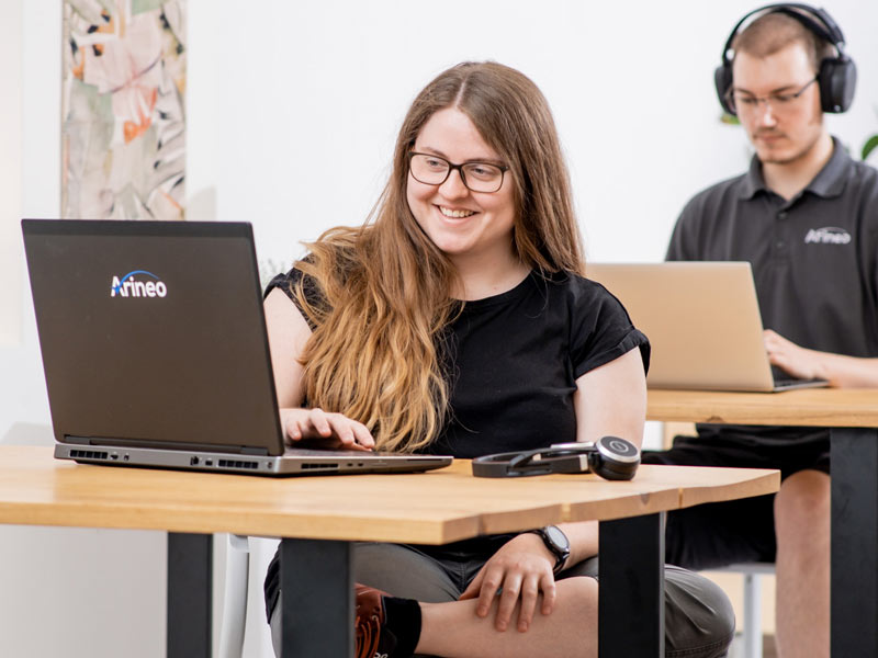 Eine Frau am Laptop optimiert das Data Management. Im Hintergrund richtet ein junger Mann einen Microsoft SQL Server für besseres Data Management ein.